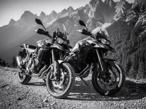 Two adventure motorcycles parked on a rocky mountain gravel trail, jagged peaks and pine forest in misty background, black and white.