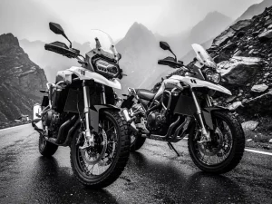 Two adventure motorcycles with knobby tires parked on a rain-slicked mountain road, misty rocky peaks looming behind them in black and white.