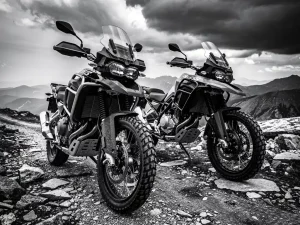 Two adventure motorcycles with knobby tires parked on a rocky mountain trail, dramatic overcast highland landscape in black and white.