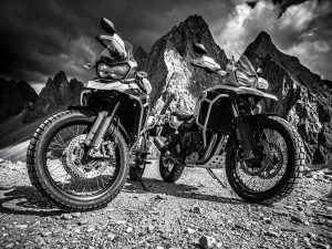 Two adventure motorcycles parked on a rugged mountain gravel trail, jagged rocky peaks behind them, dramatic storm light in stark black and white.