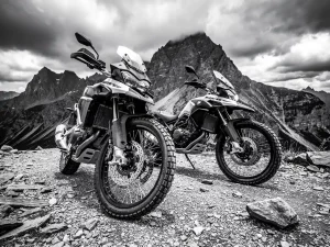 Two adventure motorcycles with knobby tires and suspension forks parked on a rocky mountain trail under a dramatic overcast sky.