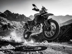 CFMOTO 450 MT adventure motorcycle airborne over rocky dirt trail, suspension compressed on landing, dust exploding beneath tires, dramatic low-angle mountain shot.