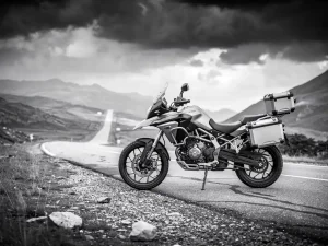 CFMOTO 450 MT adventure motorcycle parked on open asphalt road leading toward a mountain pass under dramatic overcast sky.