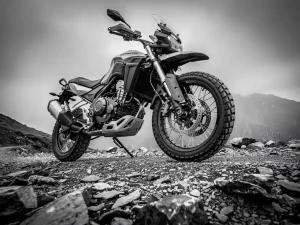 CFMOTO 450 MT adventure motorcycle on a rocky mountain trail, front forks and rear shock absorber sharp against an overcast sky, black and white.