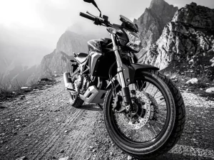 CFMOTO 450 MT adventure motorcycle on a mountain gravel road, front fork suspension in sharp focus, misty rocky peaks in background.