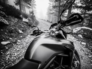 CFMOTO 450 MT adventure motorcycle on a rugged mountain gravel trail, dramatic monochrome low-angle view of fuel tank with misty pine trees behind.