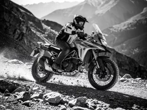 CFMOTO 450 MT adventure motorcycle cornering aggressively on rocky mountain trail, suspension compressed over jagged terrain, rider in off-road stance, monochrome.