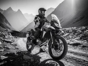 Petite rider standing flat-footed beside a CFMOTO 450 MT adventure motorcycle on a rugged mountain trail, monochrome dramatic lighting.