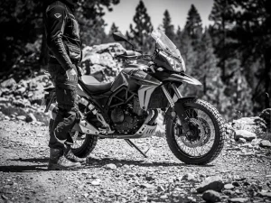 Rider standing beside a CFMOTO 450 MT adventure motorcycle on a gravel trail, demonstrating seat height reach, pine forest in background.