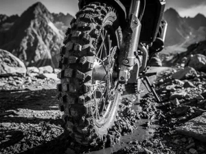 CFMOTO 450 MT knobby adventure tire tread pressed into wet rocky mountain trail mud, low-angle close-up in high-contrast black and white.