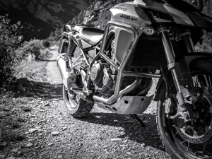 CFMOTO 450 MT adventure motorcycle with steel crash bars on a gravel mountain trail, dramatic side lighting highlighting protective engine frame in monochrome.