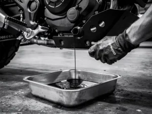 Mechanic draining dark engine oil from a CFMOTO 450 MT motorcycle into a steel drain pan, skid plate and frame visible above.