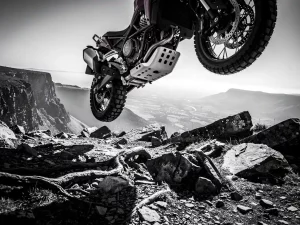 CFMOTO 450 MT adventure motorcycle airborne over rocky mountain trail, undercarriage and suspension components visible above jagged stones in misty highland terrain.