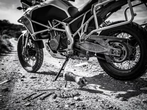 Hands bolting a steel luggage rack onto a CFMOTO 450 MT adventure motorcycle, wrenches on gravel trail, black and white photography.