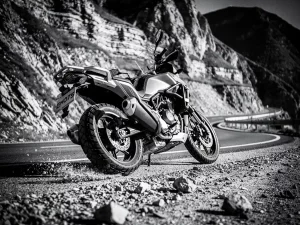 CFMOTO 450 MT adventure motorcycle carving a mountain curve, rear suspension compressed, gravel-strewn asphalt, rocky cliffs behind, dramatic grayscale sidelight.