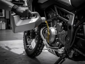 Gloved hand pouring motor oil into a CFMOTO 450 MT engine, adventure motorcycle with knobby tires visible in workshop background.