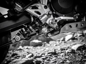 CFMOTO 450 MT rear shock absorber and linkage system in close-up, precision-machined suspension components above a rocky mountain trail.