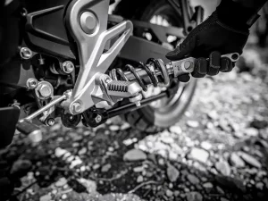 Gloved mechanic adjusting preload collar with spanner wrench on CFMOTO 450 MT rear suspension linkage, gravel trail background.