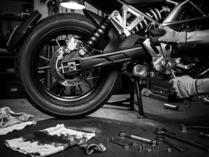 CFMOTO 450 MT adventure motorcycle on center stand with rear shock absorber exposed, mechanic adjusting suspension linkage with wrench in black and white workshop.