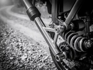 CFMOTO 450 MT front fork and rear shock absorber close-up, polished steel suspension linkage with matte rubber seals against blurred gravel trail.