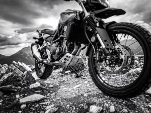 CFMOTO 450 MT adventure motorcycle suspension and front forks in sharp detail on a rocky mountain trail, dramatic grayscale low-angle shot.