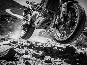 CFMOTO 450 MT suspension system close-up, front fork and rear shock absorber in sharp focus above jagged rocky trail, dramatic low-angle grayscale shot.