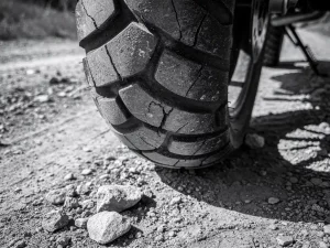 Worn CFMOTO 450 MT motorcycle tire on dirt trail, showing cracked and flattened tread grooves at contact patch with loose gravel nearby.