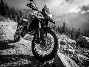 CFMOTO Ibex 450 adventure bike on a rocky mountain trail, low-angle view of handlebars and suspension forks, pine forest and misty peaks behind.