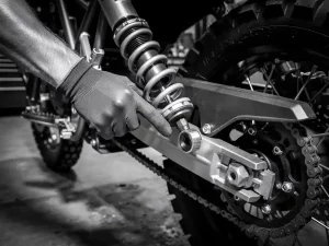 Gloved mechanic's hand adjusting rear shock absorber preload collar on a CFMOTO Ibex 450, knobby tire and frame visible behind.