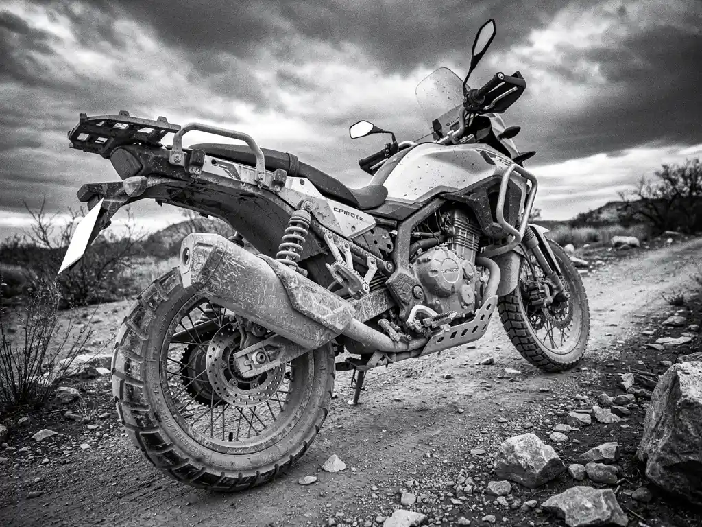 CFMOTO 450 MT adventure bike on a dirt trail, mud-caked tires and scratched bodywork after 30,000 km, black and white, low angle.