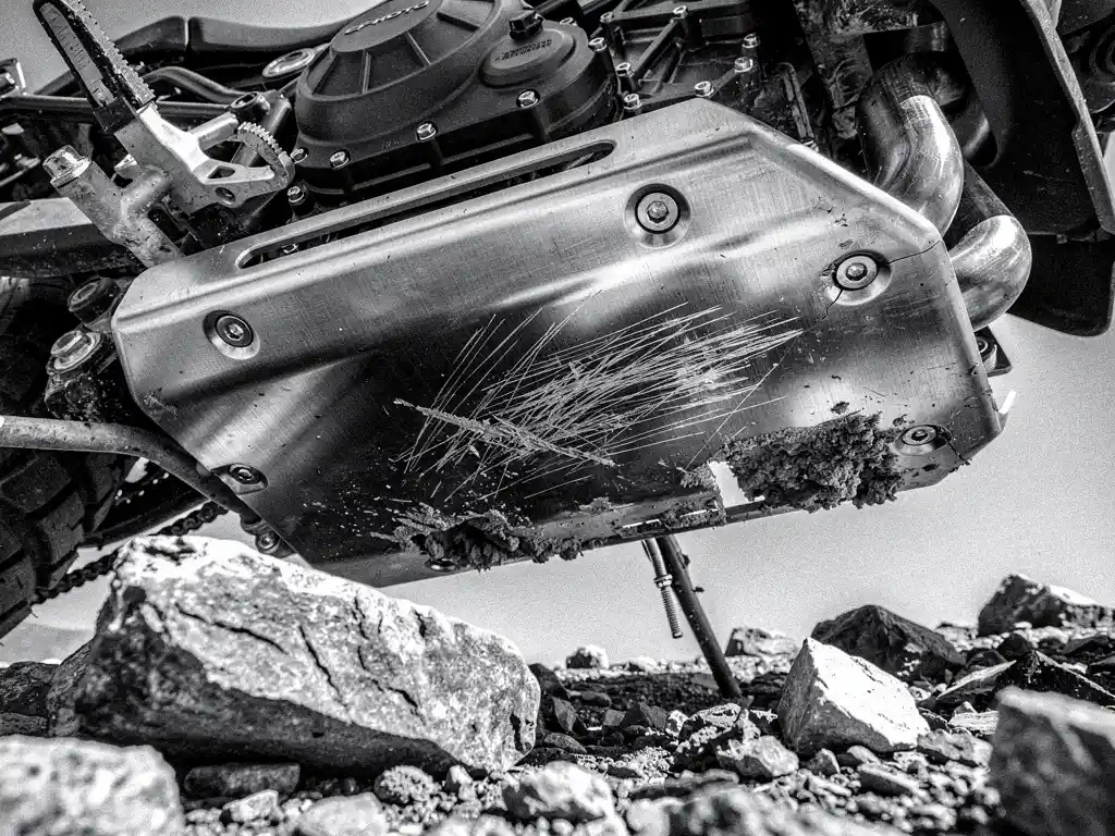 Steel skid plate on CFMoto 800 MT adventure motorcycle, scratched and mud-covered, rocky terrain visible below in high-contrast black and white.