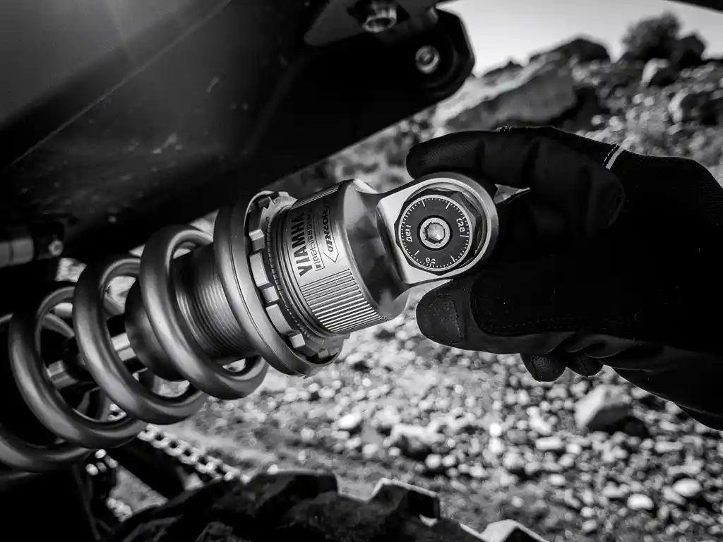 Gloved hand adjusting compression damping on a Yamaha Ténéré 700 rear shock absorber, rocky trail visible in background.