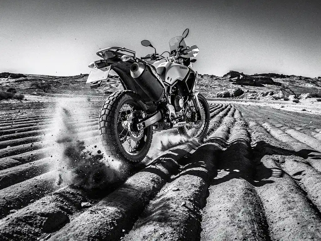 Yamaha Ténéré 700 adventure motorcycle airborne over washboard dirt track, rear suspension compressed, dust frozen mid-air, arid landscape in black and white.